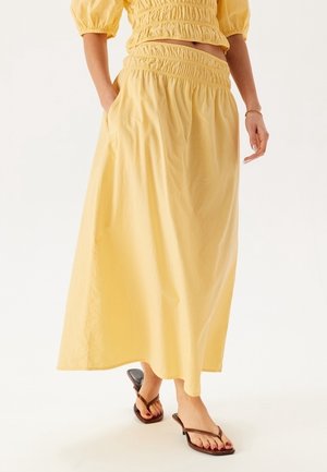 Mujer con un top corto amarillo fruncido y falda a juego hasta el tobillo con bolsillos, combinados con sandalias marrones de tacón.