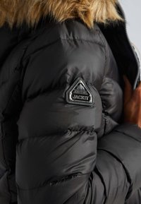 Zwarte pufferjack met een glanzende, gewatteerde textuur, voorzien van een driehoekige zwarte patch met zilveren letters "JACKIT" en een met bont gevoerde capuchon.