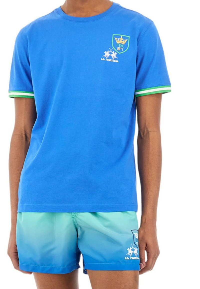 Camiseta de algodón azul con mangas a rayas verdes y logo. Combinada con pantalones cortos de baño con degradado de azul a verde que presentan un estampado.