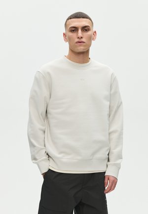 Mladić s kratkom kosom, odjeven u običnu svijetlo bež majicu dugih rukava i crne hlače, stoji na svijetloj neutralnoj pozadini.