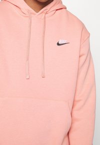 Sudadera Nike de color rosa claro con bolsillo tipo canguro, cordones y un pequeño logo bordado en blanco y negro en el pecho. Textura de tela suave.