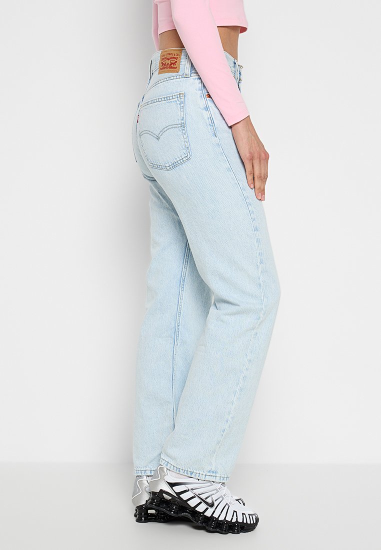 Persona vestida con jeans de tiro alto de color azul claro, camiseta de manga larga rosa y zapatillas Nike blancas, de pie contra un fondo claro y liso.