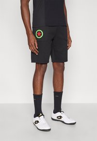 Puha anyagból készült fekete rövidnadrág, a bal zsebénél zöld és piros kör alakú logóval, fehér sportcipővel és sötét zoknival párosítva.