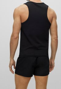 Man som bär en svart ärmlös linne och svarta träningsshorts, står med ryggen vänd mot kameran mot en enkel ljus bakgrund.