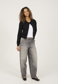 Vrouw met krullend haar draagt een zwarte cardigan met knopen over een witte top, brede grijze jeans en zwarte platte schoenen, staand tegen een effen achtergrond.