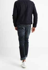 Granatowa kurtka o teksturowanej tkaninie, dopasowana w talii; zestawiona z ciemnymi dżinsami i białymi trampkami, widok z tyłu.