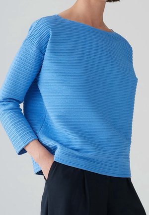Person trägt ein strukturiertes hellblaues Langarmshirt mit horizontalen Rillen und schwarze Hose, Hand lässig in der Tasche.