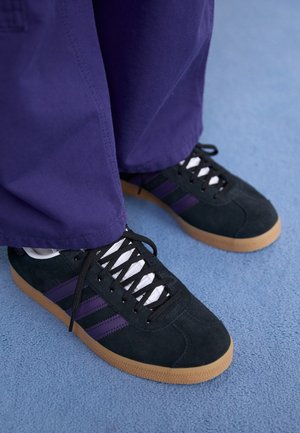 Black suede sneakers with purple stripes, white laces, and gum soles. Worn with loose purple pants, set against a blue background.