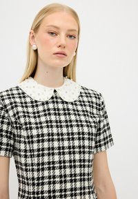 Schwarz-weißer kariertes Kleid mit strukturiertem Stoff und einem weißen, mit Perlen besetzten Kragen. Kurze Ärmel und ein taillierter Schnitt.