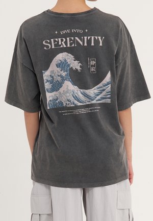 Persoon draagt een donkergrijs t-shirt met een grote gestileerde oceaangolf en de tekst "Duik in Sereniteit" op de achterkant.