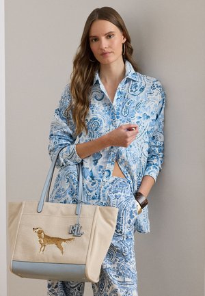 Γυναίκα με μακριά, κυματιστά μαλλιά, που φορά μπλε πουκάμισο και παντελόνι με σχέδιο παϊδάρι, κρατώντας μπεζ τσάντα ώμου με κέντημα σκύλου και γοητεία με το λογότυπο RL.