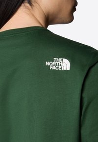 Osoba w ciemnozielonej koszuli z białym logo "The North Face" na górnej części pleców, pokazana z tyłu i z boku.