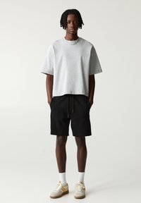 Grauer kurzärmliger Sweatshirt aus weichem Stoff, kombiniert mit schwarzen Shorts mit Seitentaschen und einem Kordelzug. Weiße Socken und Sneakers mit gelben Akzenten.