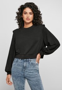 Femme aux cheveux bouclés portant un sweat-shirt à épaules rembourrées noir et un jean taille haute bleu clair, se tenant devant un fond uni.