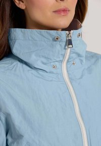 Lichtblauwe jas met een hoge kraag, volledige rits aan de voorkant en drukknoopaccenten. Kenmerkt zich door een gladde textuur en gestructureerd ontwerp.