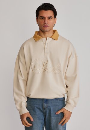 Muž s tmavými vlasmi nosí béžovú dlhú polokošeľu s hnedým golierom a vyšitým logom "Siksilk", v kombinácii s modrými džínsami.