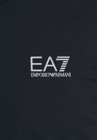 EA7 Emporio Armani bílý logo na černé látce, kombinující text s vodorovným pruhovaným designem na čísle 7.