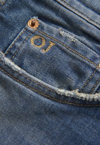 Niebieska tkanina dżinsowa z widocznymi szwami i wyblakłym detalem kieszeni. Zawiera brązowy guzik i haftowane inicjały "OJ". Występuje lekkie przetarcie.