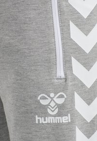 Grå sweatpants med en vit dragkedja och en stor vit logotyp. Har ett chevron-mönster på sidan, tillverkat av mjukt, strukturerat tyg.