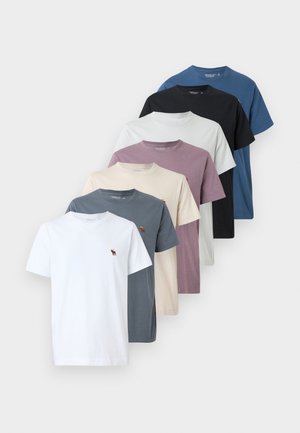 Uma coleção de sete t-shirts de gola redonda em várias cores: branco, cinza, bege, roxo, azul, preto e azul claro, com um pequeno logotipo.