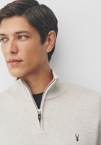 Beige, ribbed knit pullover with a high collar and quarter zipper; features a small dark emblem on the chest. Cotton blend material.