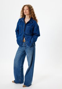Chaqueta de mezclilla con dos bolsillos en el pecho, corte oversized y color azul. Combinada con jeans de pierna ancha, tejido texturizado y detalles de costura sutiles.