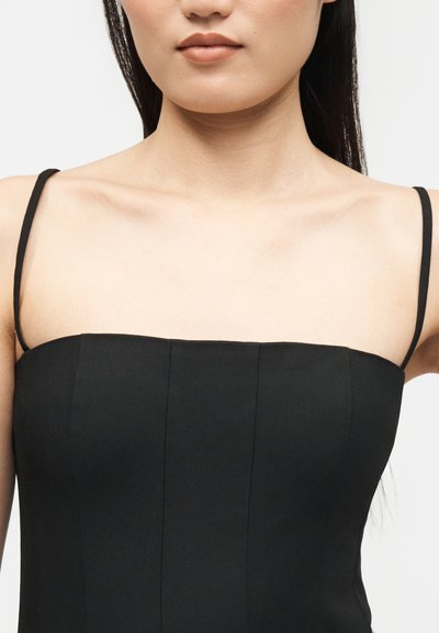 Nő fekete, pánt nélküli ruhában, vékony vállpántokkal, látható nyakkivágással és mellkas felső részével, egyszínű háttér előtt.