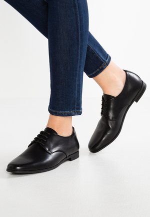 Osoba nosząca ciemnoniebieskie wąskie dżinsy i czarne skórzane eleganckie buty na sznurowadła, stojąca na białej podłodze z jedną uniesioną stopą.