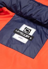 Binnenkant van een rode en marineblauwe pufferjack met een LevelUP-label. Kenmerkt zich door een gladde, glanzende textuur, vierkante patchontwerp en contrasterende kleuren.