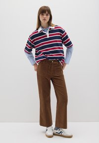 Camiseta tipo polo a rayas en navy, rojo y blanco; pantalones de pana anchos color marrón; zapatillas blancas con detalles en navy; modelo posando frente a un fondo liso.