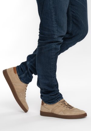 Person trägt dunkelblaue Jeans und beige Wildledersneaker mit braunen Sohlen, steht mit einem Fuß hinter dem anderen verschränkt.