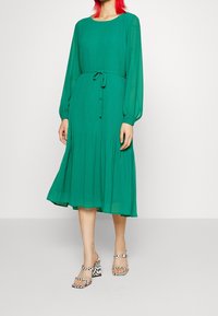 Zelené midi šaty s plisováním a dlouhými rukávy, opatřené vázáním v pase a kulatým výstřihem. V páru s černobílými kostkovanými sandály s blokovým podpatkem.