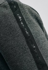 Szary sportowy top z fakturowanej tkaniny oraz czarną wstążką z nazwą marki wzdłuż boku, wyszytą w odblaskowym wykończeniu.