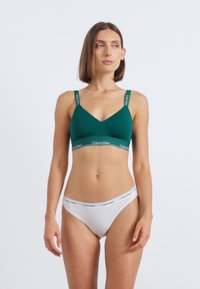 Egy nő áll, zöld Calvin Klein melltartóban és fehér Calvin Klein alsóban, kissé oldalra nézve, egy egyszínű háttér előtt.