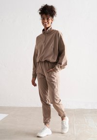Světle hnědý teplákový komplet vyrobený z hladké tkaniny, s límcem na půl zip, elastickým pasem a manžetami, doplněný o bílé tenisky.