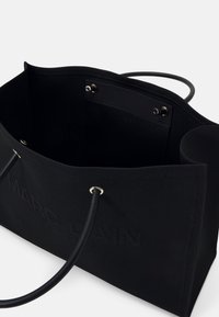 Sac cabas rectangulaire noir avec deux poignées rondes, poche intérieure visible avec boutons-pression et nom de marque subtilement embossé à l’extérieur.