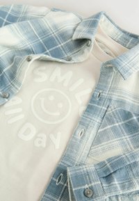 Witte shirt met een glimlachend gezicht en de tekst "SMILE All Day" onder een open blauw-witte geruite blouse.