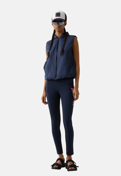 Kvinde med fletninger iført navyblå, ærmeløs polstret vest, navyblå leggings med pink logo, sorte sandaler, hvid og sort kasket samt mørke solbriller.