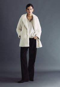 Kobieta stoi, ubrana w biały płaszcz z futra syntetycznego, beżowy dziany sweter, ciemne szerokie spodnie oraz czarne buty, z złotymi kolczykami-kołami na tle szarego tła.