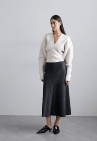 Balta adīta džemperis ar V veida kakla izgriezumu un apjomīgām piedurknēm, kombinēts ar tumši pelēku A līnijas midi svārku un melniem šņorējamajiem apaviem.