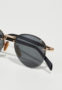Ochelari de soare rotunzi cu lentile întunecate, accente metalice aurii și brațe din plastic negru cu textură pentru aderență. Branding vizibil pe interior.