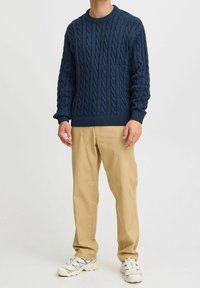 Pull en maille torsadée bleu marine avec encolure et poignets côtelés, associé à un pantalon kaki et des baskets de couleur claire. Design classique et texturé.