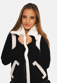 Jeune femme aux longs cheveux portant une veste noire bordée de moumoute blanche, tenant le col et regardant la caméra avec un léger sourire.