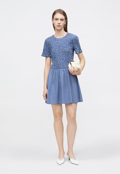 Femme portant une robe courte à manches courtes bleu texturé avec une jupe évasée, tenant une petite pochette blanche et portant des mules blanches à talons.