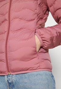 Prošívaná bunda v jemně růžové barvě s vlnitým švem, zapínáním na zip a bočními kapsami. Vybavena elastickými manžetami a spodním lemem pro těsné padnutí.