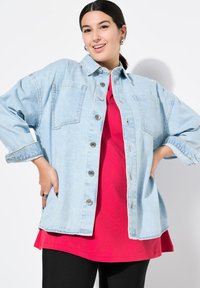 Veste en denim oversized bleu clair avec des boutons argentés, deux poches poitrine et des manches retroussées, superposée sur une chemise rouge et un pantalon noir.
