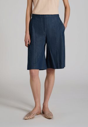 Frau trägt knieumspielende dunkelblaue Shorts mit weitem Bein und Seitentaschen, beige ärmelloses Oberteil und beige gewebte Slip-ons.