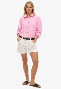 Lichtroze lange mouwen shirt met knopen, gecombineerd met witte denim shorts met rafelige zomen en een zwarte riem, gedragen met zwarte slide sandalen.