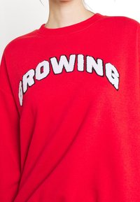 Červená mikina s kulatým výstřihem, na které jsou bílé a černé vyšívané písmena „BROWNING“ na hrudi. Měkká textura.