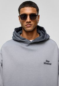Grijze hoodie van textuurstof, accentueert met een donkergrijze voering. Bevat een "Stay Moment" grafiek in zwart. Zwarte zonnebril maakt de look compleet.